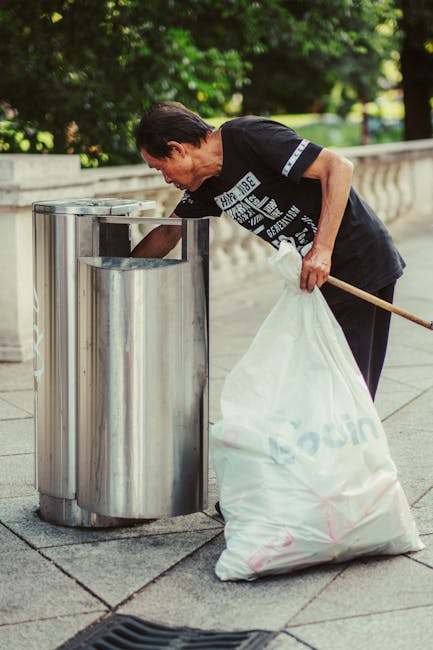 A man wearing a dark T-shirt and black trousers is outdoors, leaning over a stainless steel litter bin on a paved pathway, emptying refuse into a large white rubbish bag he holds in his left hand. The