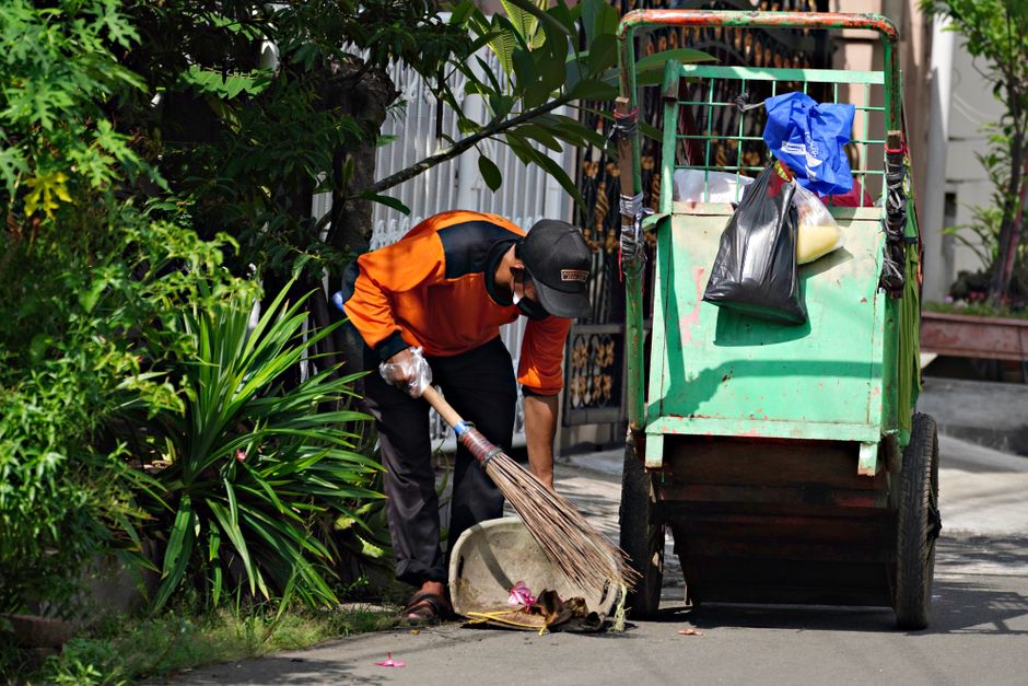 A man wearing a dark T-shirt and black trousers is outdoors, leaning over a stainless steel litter bin on a paved pathway, emptying refuse into a large white rubbish bag he holds in his left hand. The